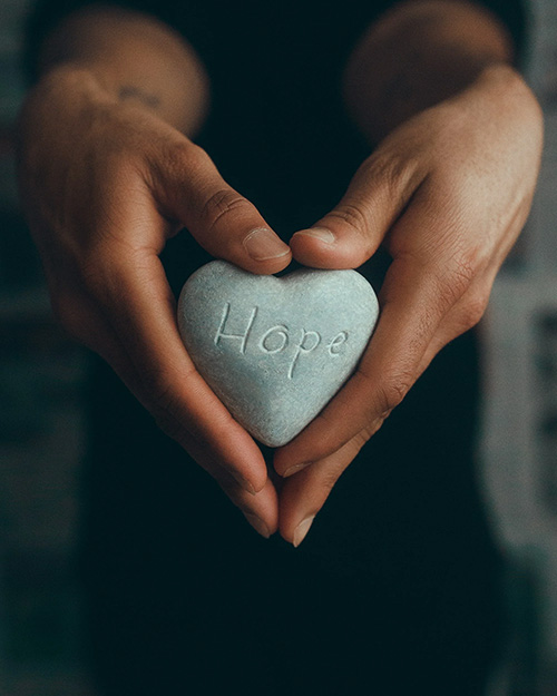 A pair of hands holds a heart-shaped stone with the word 'Hope' carved into it