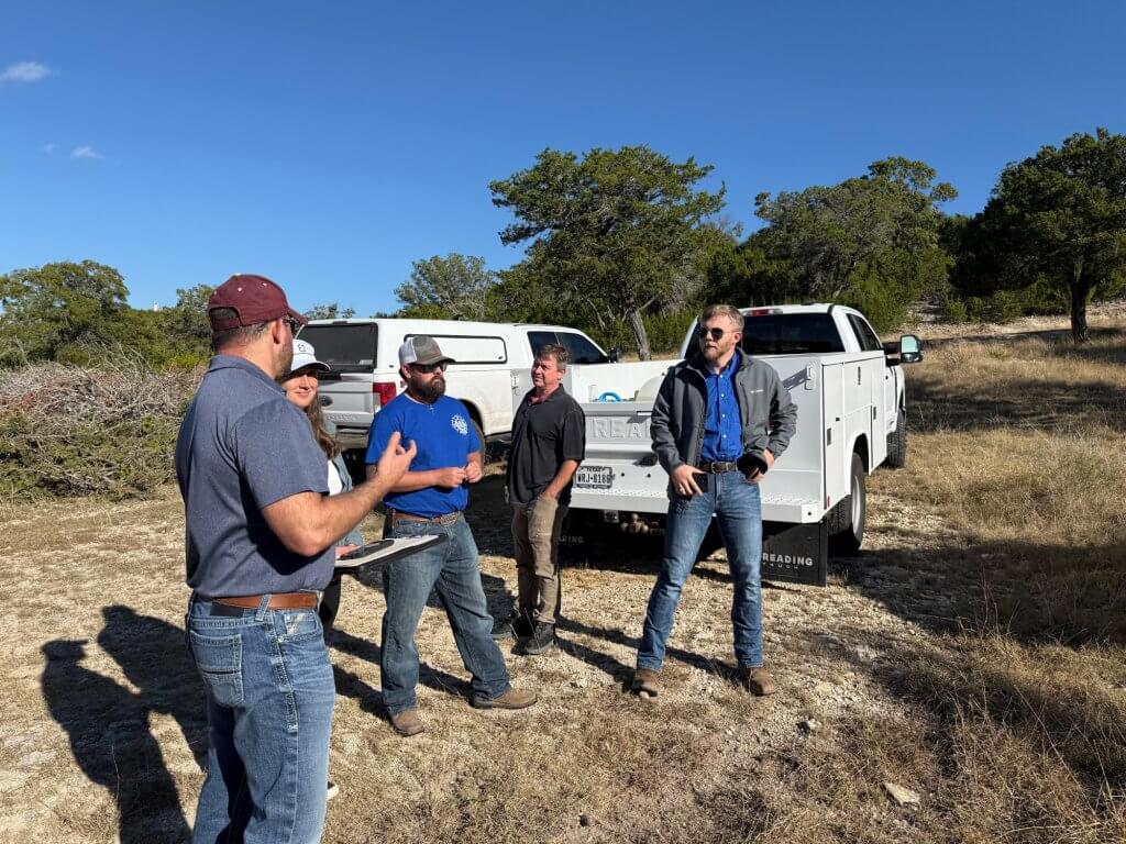 A group of six people in casual attire are gathered around a white utility truck in a dry, grassy field