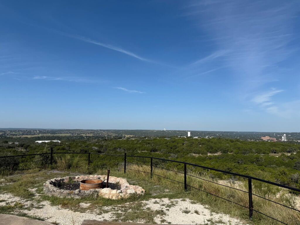 A panoramic view of a grassy hillside with a stone fire pit and a black metal fence overlooking a vast landscape