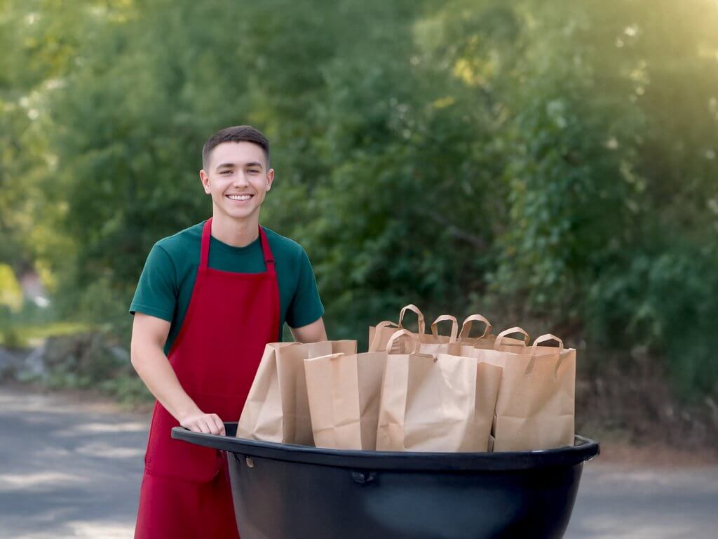 A young man in a red apron stands outdoors with a cart full of brown paper bags