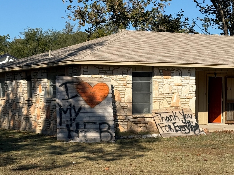 A house with a stone facade has a wooden sign with graffiti saying 'I ❤️ My HEB' and 'Thank You For Everything'