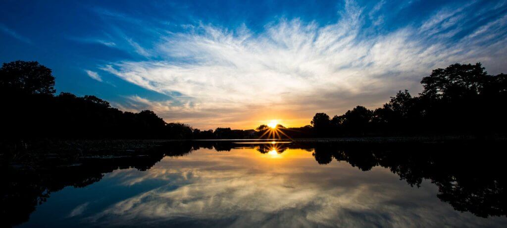 A serene sunset reflects on a calm lake, with trees silhouetted against a vibrant sky