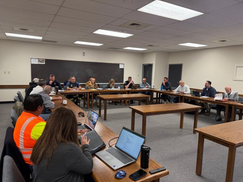 A group of people in a conference room, some wearing safety vests, are seated around tables with laptops and documents
