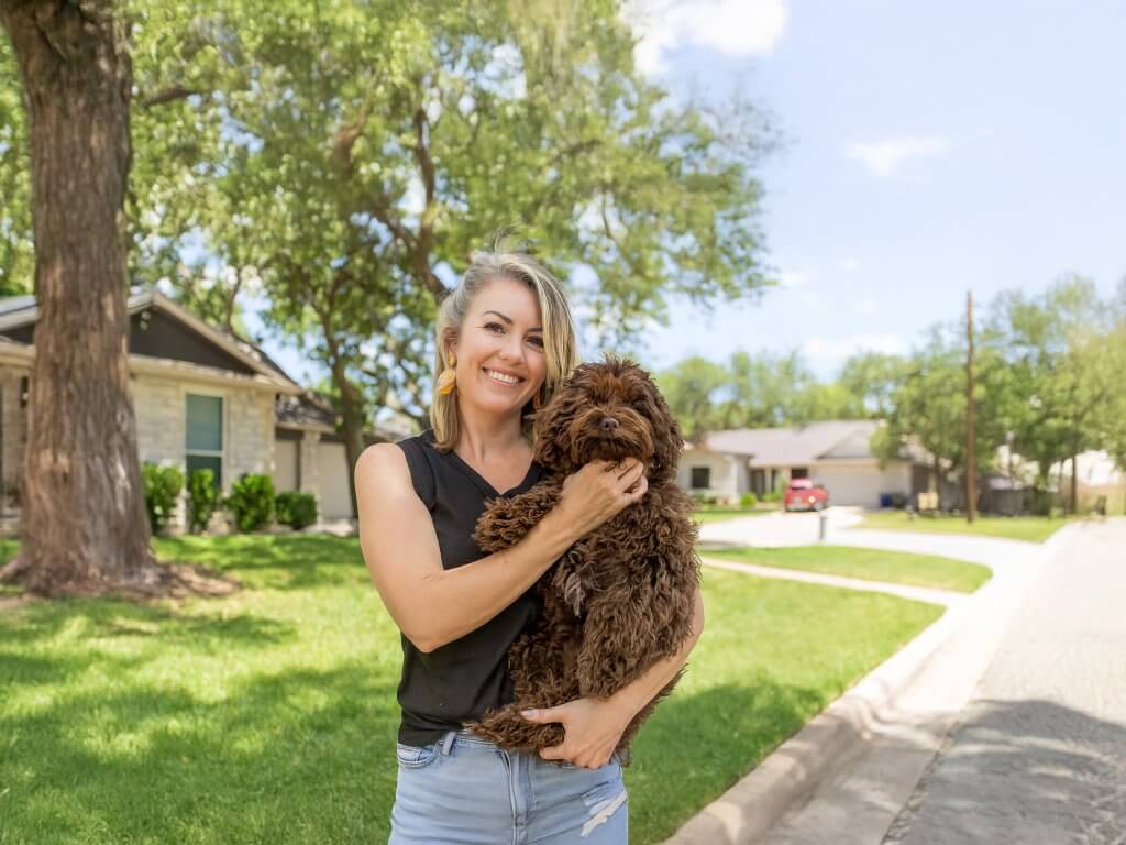 Woman with blonde hair smiles while holding a brown curly-haired dog in a residential area